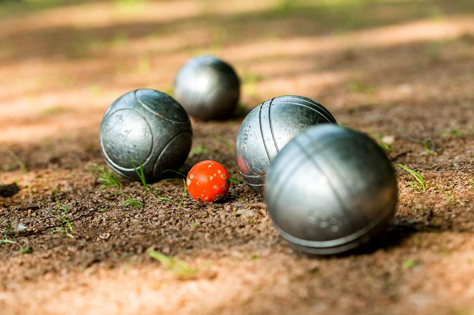 Gleise mit silbernen Boulekugeln und einer roten Kugel auf einem erdigen Boden in einem Park.