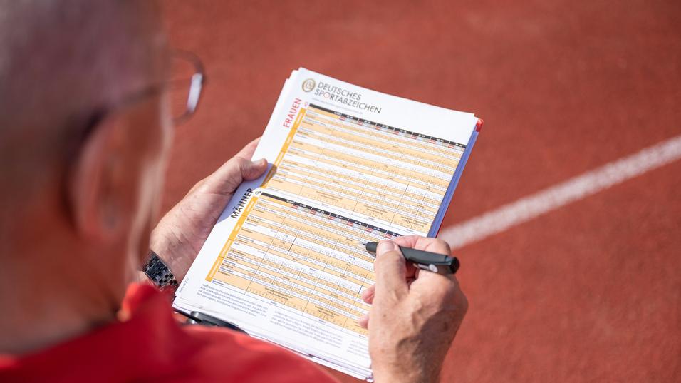 Hand eines Prüfers, der auf einem Blatt mit Sportabzeichen-Anforderungen auf einer Laufbahn notiert.
