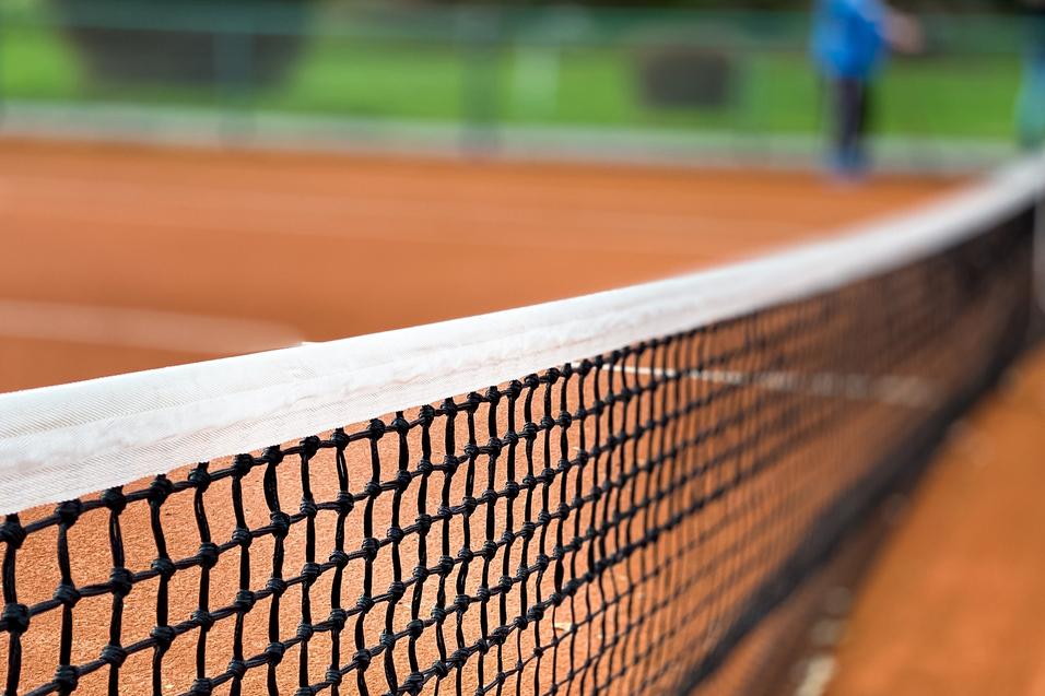 Nahaufnahme eines Tennisnetzes auf einem roten Sandplatz, Hintergrund unscharf mit grünen Bäumen.