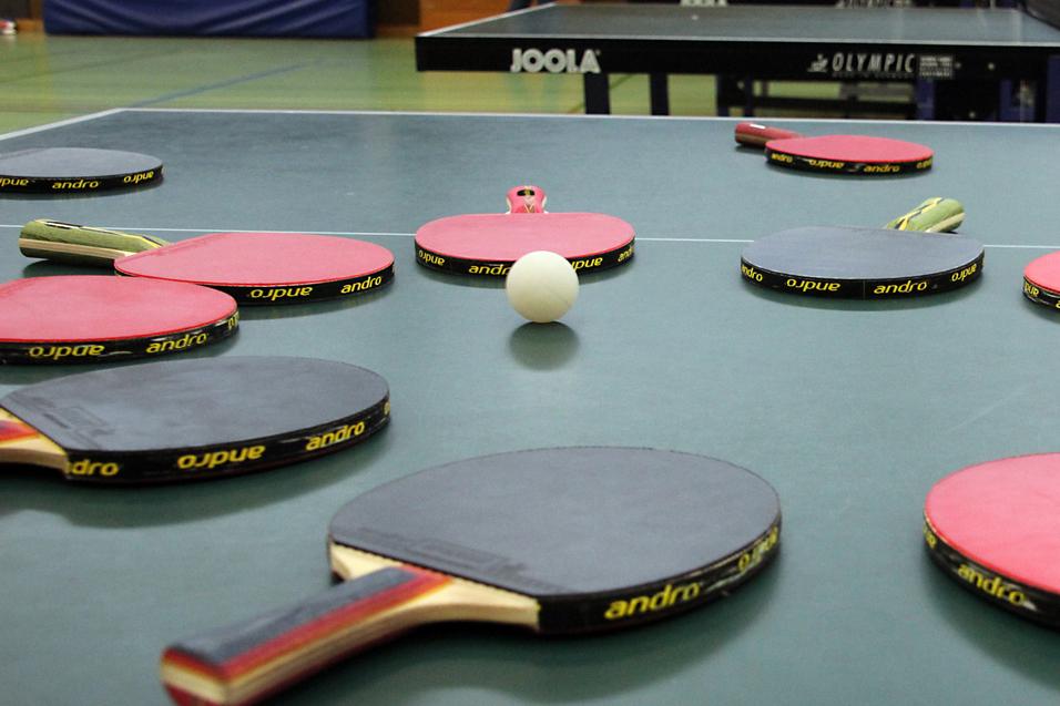 Mehrere Tischtennisschläger in verschiedenen Farben und ein Tischtennisball liegen auf einer grünen Tischtennisplatte.